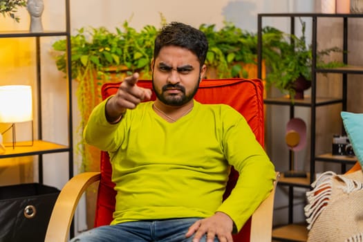 Hey you. Strict unhappy Indian man pointing hand finger to camera, looking angry, making choice, choosing guilty, noticing suspicious, accusing for mistake at home room apartment. Guy sits on chair