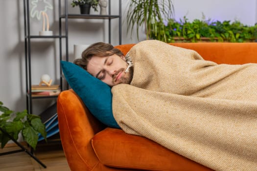 Tired young man lying down in bed taking a rest at home apartment room. Carefree Caucasian guy napping, falling asleep on comfortable orange couch with pillows. Closed his eyes enjoy daytime nap alone