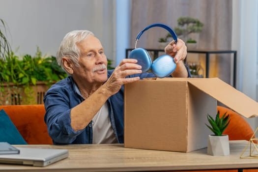 Happy senior man unpacking delivery parcel with headphones at home. Smiling satisfied grandfather shopper, online shop customer opening cardboard box receiving purchase gift by fast postal shipping