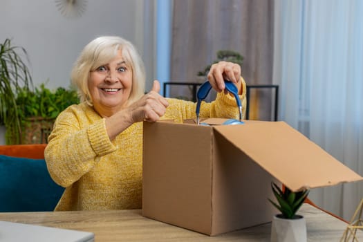 Happy senior woman unpacking delivery parcel with headphones at home. Smiling excited grandmother shopper, online shop customer opening cardboard box receiving purchase gift by fast postal shipping