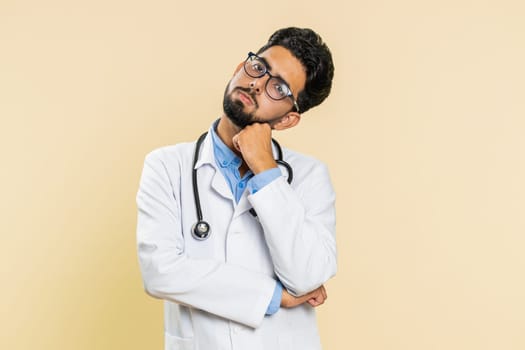 Sad Indian young doctor cardiologist man tired bored exhausted indifferent expression, not interested in communication talk, displeasure. Arabian apothecary pharmacy guy isolated on beige background
