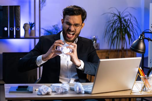 Angry furious Indian man working at office throwing crumpled paper, having nervous breakdown at work, migraine headache, stress management, mental distress problems, losing temper, reaction on failure