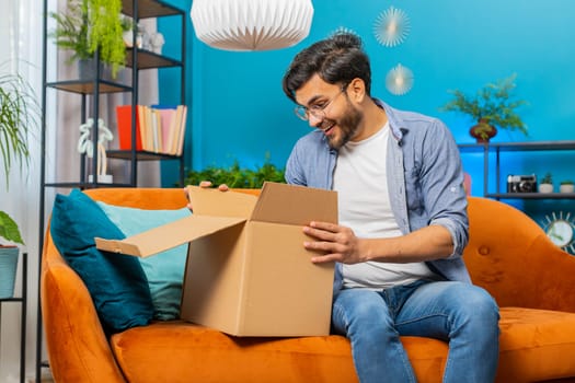 Happy Indian young man unpacking delivery parcel sits at home. Smiling satisfied Arabian Hindu guy shopper online shop customer opening cardboard box receive purchase gift by fast postal shipping