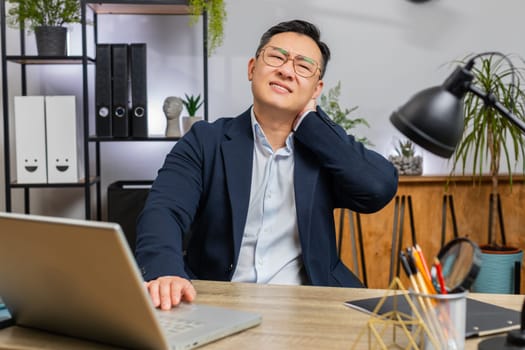 Tired Asian middle-aged businessman suffers from spine neck muscle pain at home office workplace working on laptop. Overworking. Freelancer Chinese man in suit attempt to relieve pain by massaging