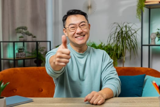 Like. Happy excited Asian man looking approvingly at camera showing thumbs up like sign positive something good great news positive feedback. Middle-aged Chinese guy sits on couch at home living room