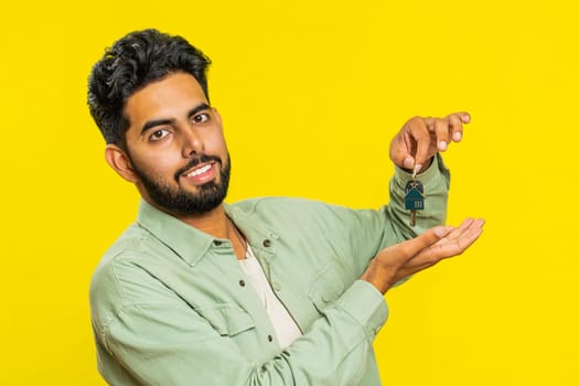 Indian young man real estate agent lifting hand showing the keys of new home house apartment, buying or renting property, mortgage loan, insurance. Arabian guy isolated on yellow studio background