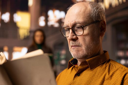 Retired man enjoys book reading at a public library and for leisure activity, feeling relaxed and increasing his knowledge during retirement. Happy calm person with eyeglasses, recreation.