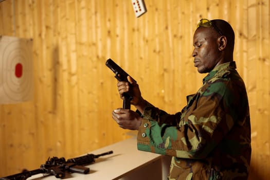 Army soldier in shooting range reloading handgun magazine, preparing to shoot targets. African american man at firing range loading pistol during marksmanship training program