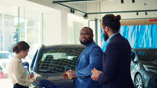 Clients arriving in car dealership, being greeted by friendly car salesman ready to showcase available vehicles to clients. Customers walking in showroom, being welcomed by agent, camera A