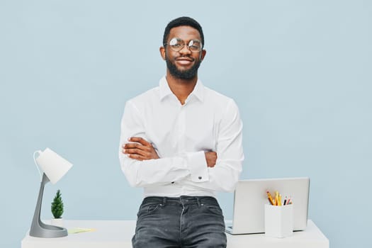 Friendly young man with glasses standing at sleek office desk with crossed arms, smiling, contemporary workspace with laptop, white lamp, and stationery, professional atmosphere, light blue backdrop, positive mood