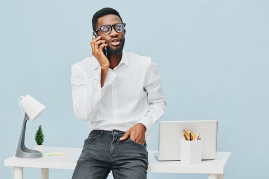 Confident young man in formal clothing engaging in a phone call inside a contemporary office environment, standing near a desk with a laptop and stationery, conveyance of business communication, professional atmosphere, workspace, office lifestyle