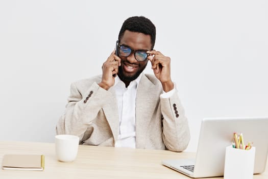 Cheerful man with dark skin, curly hair, and glasses, sitting at a desk with a laptop, sponge holder, and cup, smiling and adjusting glasses, minimalist bright office setup, casual business attire, positive professional mood, concept: workplace confidence.