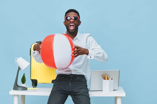 Joyful man wearing sunglasses and a white shirt holding a big beach ball, standing at a desk with office supplies and a plant, against a vivid blue background. Concept: work-life balance, summer fun, relaxation, playful mood, lifestyle photography, studio image, energetic vibe.