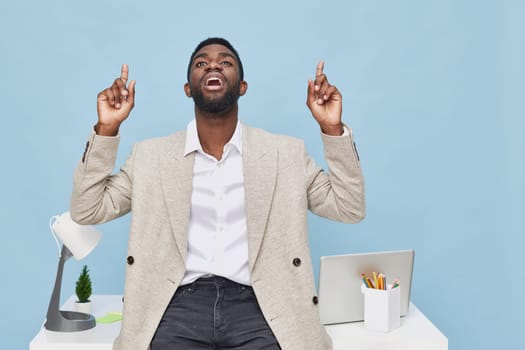 Happy and motivated man of African origin, dressed in smart casual, celebrating achievement or idea, sitting at a tidy workplace with desk accessories and computer, on a vibrant blue background. Concept: workplace positivity.