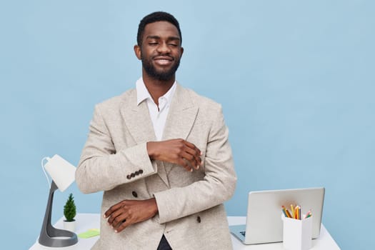 Portrait of a confident young African man in a stylish beige blazer and white shirt, smiling and looking at the camera in a modern office environment with a laptop, stationery, and a minimal pine plant, emphasizing professionalism and success. Concept: professional confidence