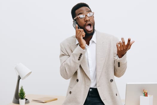 Excited young African American man in beige blazer engaged in conversation on mobile phone, gesturing with his hand, standing at clean office workspace with white background, conveying surprise, communication challenge, and workplace stress.