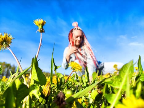A person with bright red and white braids interacts playfully with dandelions in a sunny field against a blue sky.