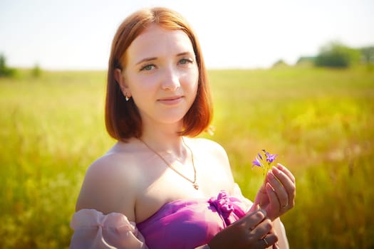 A young woman stands in a vibrant field, wearing a purple dress and holding delicate wildflowers while enjoying the sunny day.