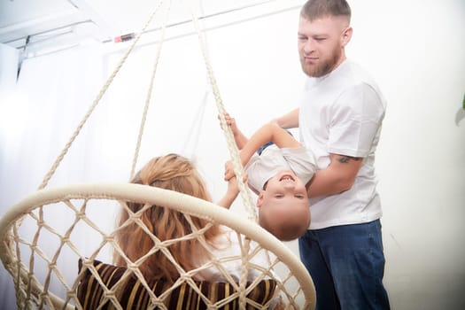 Father lifts his smiling child while the mother watches happily from a swinging chair, creating joyful family memories in a bright room.