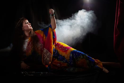A mature woman styled in a colorful sari poses elegantly in a dim setting, highlighting her beauty and the rich cultural attire she wears.