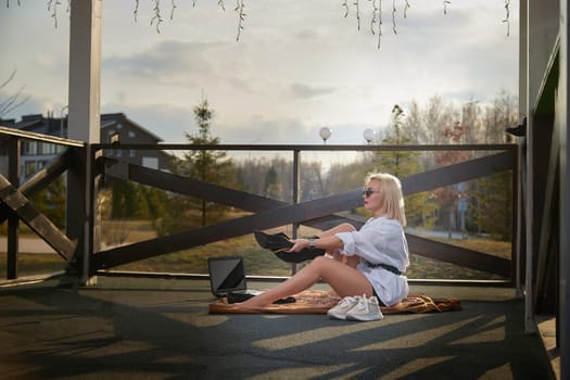 A young woman adjusts her shoes while seated on a cozy balcony, preparing for outdoor work on a bright, sunny day.