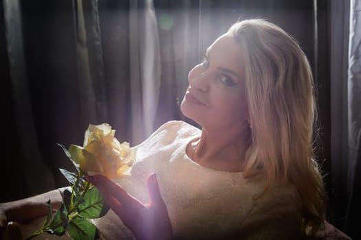 A middle-aged woman poses gracefully with a yellow rose in a cozy indoor setting, reflecting her blogging style.