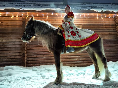 A Russian girl in traditional attire enjoys a photoshoot with a horse amidst winter decorations for Maslenitsa.