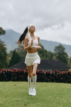 Energetic young woman wearing a white athletic outfit running outdoors, representing a healthy lifestyle, fitness, and determination in a vibrant green environment
