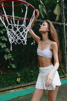 A sporty female model in a white crop top and skirt, showing determination as she reaches for the basketball hoop in a lush, vibrant setting, ideal for health and fitness themes