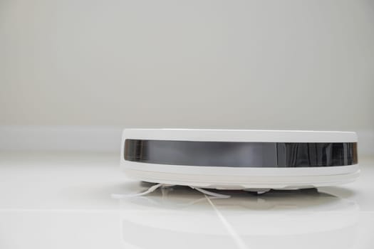White automatic floor cleaner with side brush on tile flooring, featuring front black sensor strip. Robot vacuum cleaner efficiently cleaning floor, illustrating smart home technology for dust removal