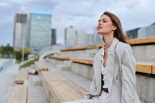 Thoughtful woman in light gray business suit sitting on outdoor bleachers with modern city skyline blurred, expressing calm confidence and autumn atmosphere, business concept