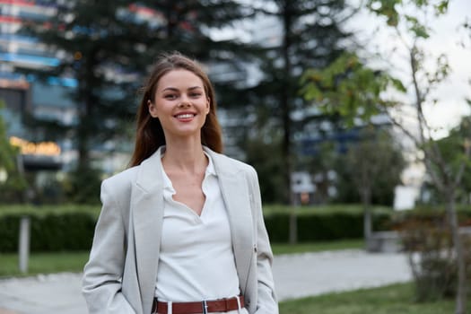 A confident woman in stylish business attire smiles warmly while standing in an urban park setting, reflecting professionalism and a positive attitude in her environment.