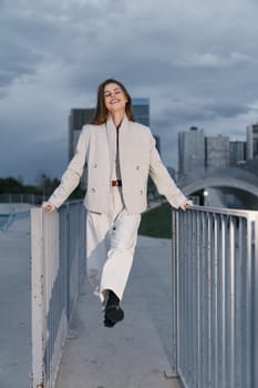 confident young woman in beige suit walking outdoors in urban city environment smiling with joy against modern buildings and cloudy sky, fashion business concept