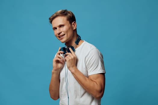 Young cheerful man with headphones around his neck smiling at camera, wearing casual t-shirt, relaxed and happy, photo taken studio setting with blue background, good for music and lifestyle themes. Concept: music enjoyment.