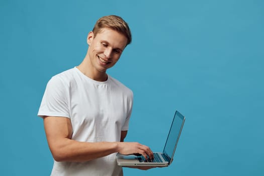 Happy young man with blonde hair in a white t-shirt working on his laptop against a solid blue backdrop. Casual and professional vibe emphasizing technology, communication, and youthful energy. Concept: digital lifestyle.