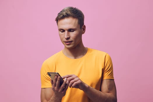 Young man in yellow t-shirt holding and interacting with a mobile phone against a pink studio background, relaxed and happy expression, modern lifestyle, technology use, communication concept, vibrant color palette