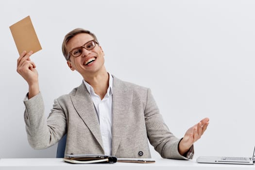 happy smiling middle aged caucasian man wearing glasses and beige blazer hold envelope sitting at table with laptop paperwork work remote freelance digital nomad concept of business communication and home office lifestyle