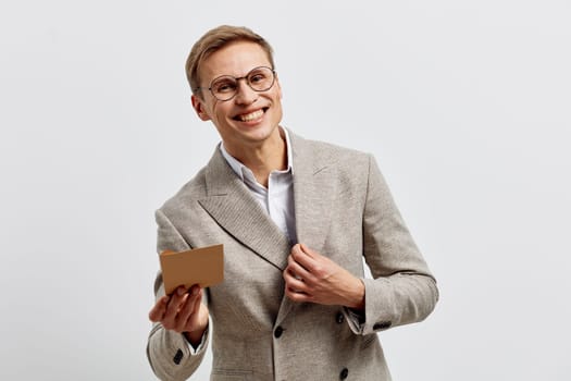 Cheerful middle-aged man with glasses dressed in a light gray blazer holding a small cardboard box, smiling happily against a plain gray background. Concept: successful delivery or gift exchange