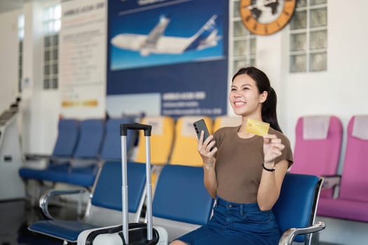 A smiling young woman at the airport proudly showcasing her phone and credit card, radiating enthusiasm for her upcoming travel.