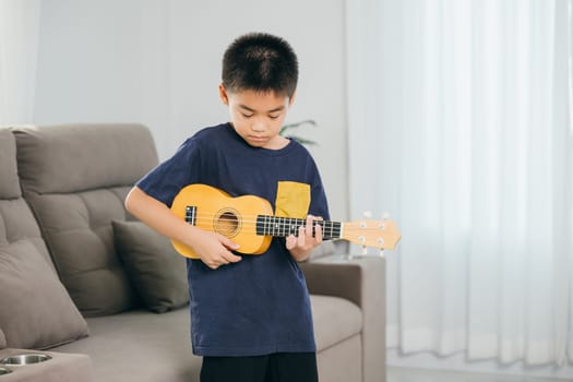 Asian kid boy stands in a living room, playing a ukulele with enthusiasm. The bright space highlights his love for music and creativity, blending a homey vibe with a playful musical moment.