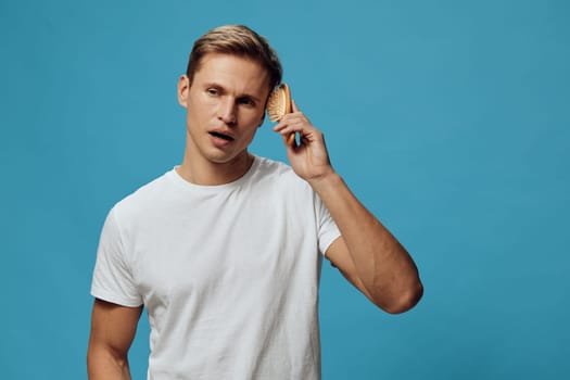 Peaceful young man with short brown hair and light skin, dressed in a simple white t-shirt, holds a hairbrush near his ear, showing a surprised or confused expression while standing against a vibrant blue backdrop. Studio shot. Concept: daily routine