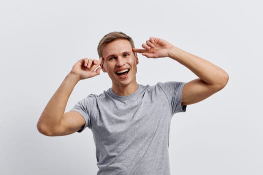 Happy young man with light brown hair in casual gray t-shirt smiling and playfully holding his ears, looking at camera against a plain white studio background. Positive emotion, joy, carefree mood