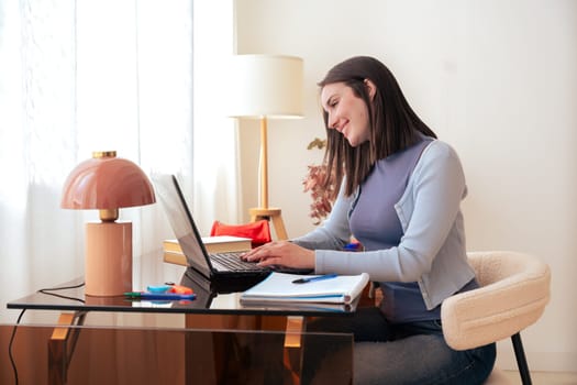 Caucasian smiling student studying at home, writing in notebook while using laptop. Modern workspace with stylish decor, lamp, and stationery. Education, learning, productivity, and concentration.