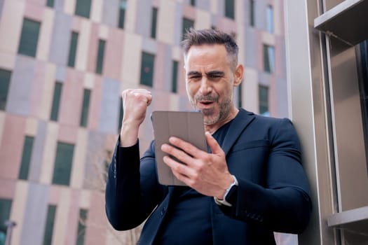 Confident businessman celebrates winning an online bet, holding a tablet and raising his fist. He smiles outdoors in a city setting. Concept of success, digital lifestyle, and modern finance.