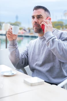 Man with short dark hair and beard drinks coffee while talking on his smartphone.He wears a casual gray sweatshirt and stands outdoors near the water,looking focused in a modern urban setting Vertical