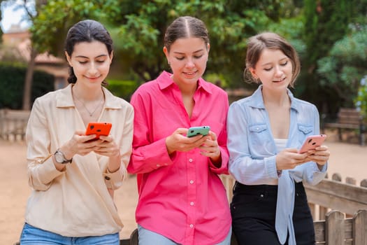 Three young Caucasian women on a wooden bench in a green park,smiling while using smartphones.they look happy and engaged,enjoying a casual outdoor moment with modern devices and social media apps