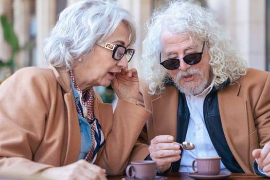 Caucasian senior couple enjoying coffee at an outdoor cafe. The woman speaks closely to the man, creating an intimate and thoughtful moment. Concept of connection, trust, and shared experiences.