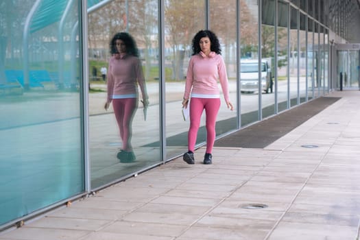 Caucasian athletic woman in pink sportswear walking beside a modern glass wall. She holds a notebook and pen, with clear copy space on the left. Urban setting, casual and active lifestyle.Copy space