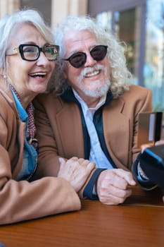 Joyful caucasian senior couple at an outdoor cafe making a card payment. Both wearing trendy outfits and sunglasses. Ideal for retirement, finance, lifestyle, and senior couple relationship concepts.