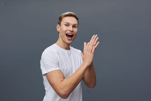 Happy cheerful man in casual white t-shirt showing excitement and joy, facing camera with hands together, on gray studio background. Concept: positive emotion and energy